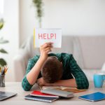 Person sitting at desk studying with head on the desk holding up a HELP sign