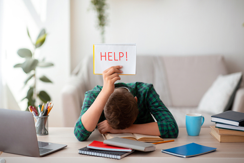 Person sitting at desk studying with head on the desk holding up a HELP sign